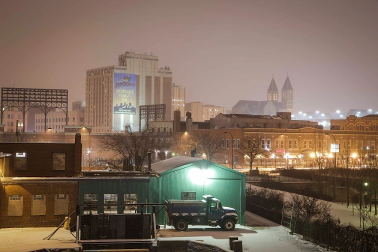 Part of the Akron skyline appears beneath a gray night sky, while in front of it a dump truck sits in a parking lot lightly coated with snow.